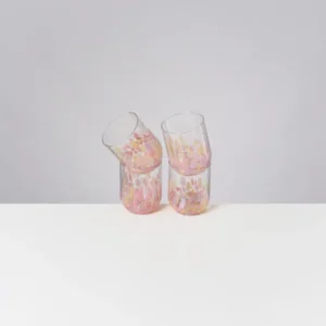Vier bunte Gläser mit pastellfarbenem Muster stehen auf einem weißen Tisch vor grauem Hintergrund.