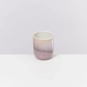 Keramiktasse mit einem Farbverlauf von Creme zu Lila, auf einer weißen Oberfläche vor grauem Hintergrund.
